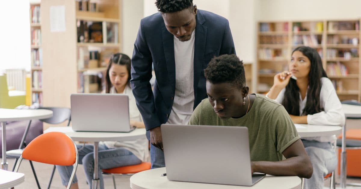 Students working on laptops in a classroom while a tutor helps one of them